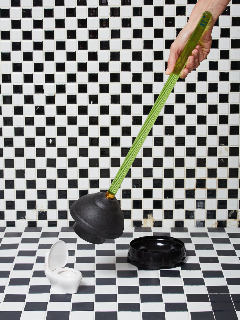 A hand demonstrates the green plunger held up against a tiny toilet model.