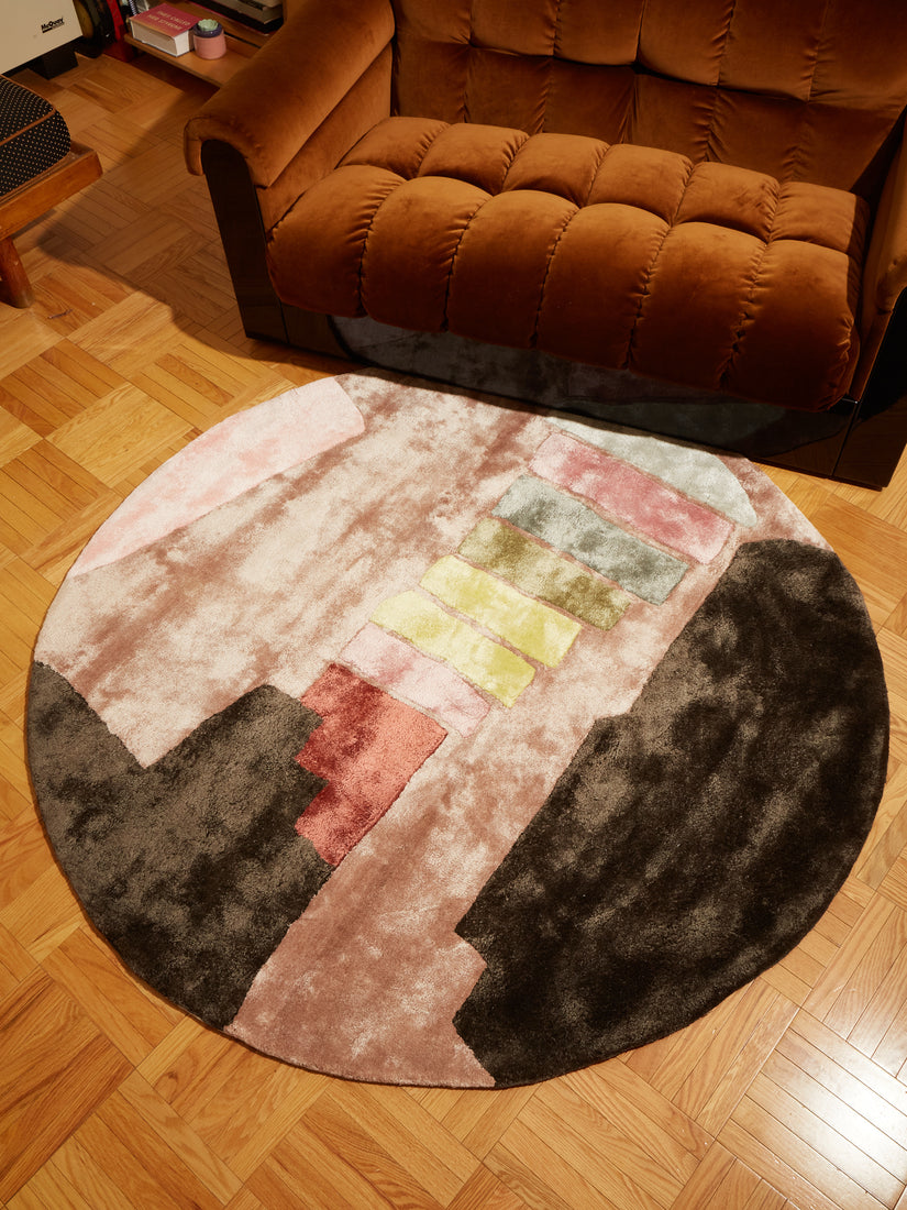 5x5 Round The Dessert Rug by Cold Picnic on a wooden floor next to a brown velvet couch.