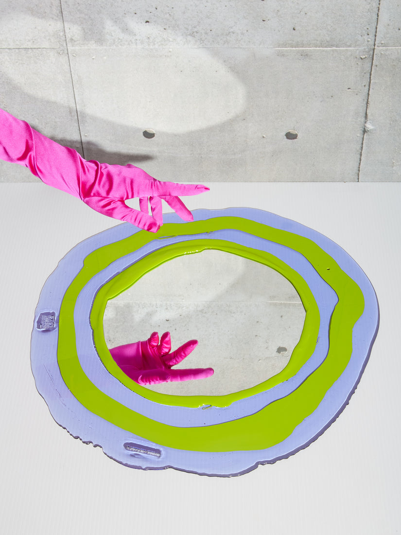A hand in a pink glove hovers over the Round Mirror in lilac and lime showing the reflection of the mirror.