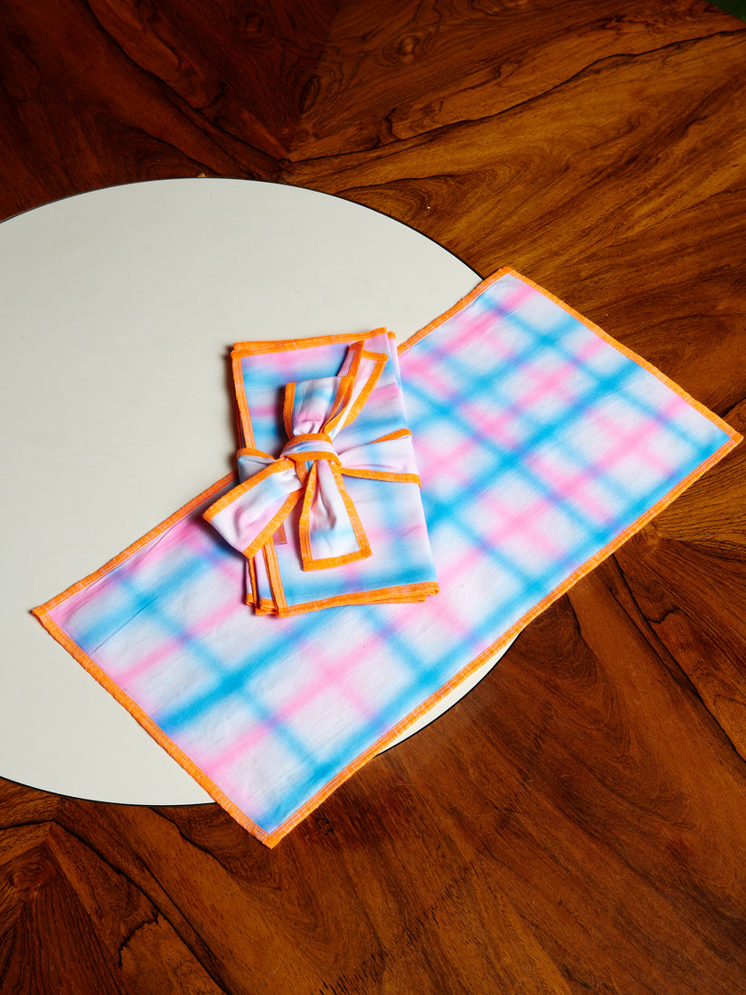 Neon orange, pink, and blue grid airbrushed napkins, one layed out against a dark wooden dining table, and one folded and wrapped in a ribbon of matching material.