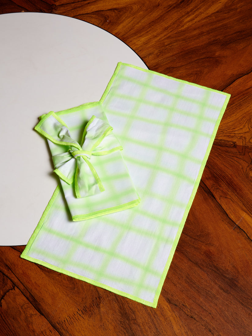 Neon green grid airbrushed napkins, one layed out against a dark wooden dining table, and one folded and wrapped in a ribbon of matching material.