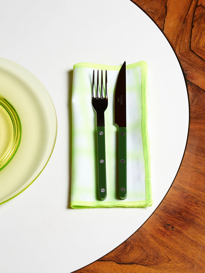 A green fork and knife by Sabre sit atop a green and white napkin by Hotel Elma just to the right of a transparent green dinner plate by Mosser.