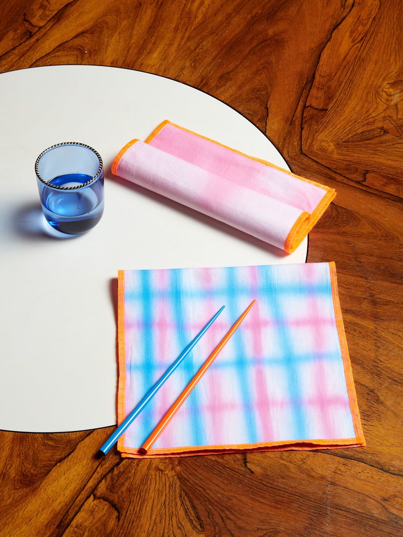 Orange and blue chopsticks sit across a folded pink, blue, and orange Hotel Elma dinner napkin. Above that sits a rolled pink and orange napkin, next to a blue striped rim glass by Fredericks and Mae.