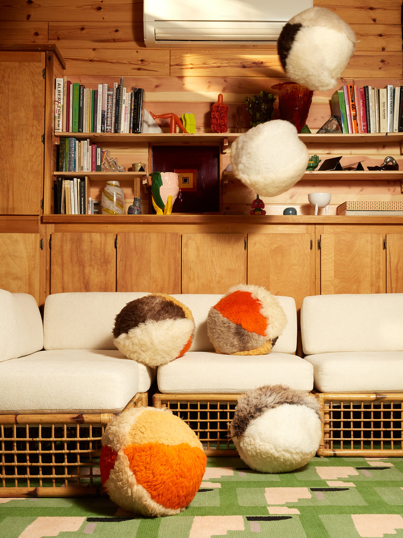 Sheepskin Orb Pillows by Casa Ahorita being resting on the floor, atop, and flying over a white and rattan couch in a green carpeted room.