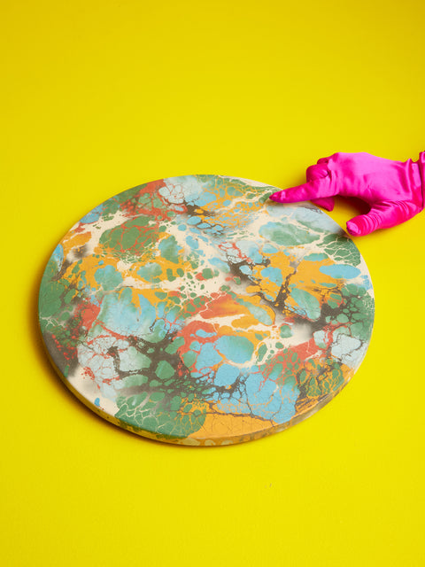 A pink satin gloved hand points at the edge of a round concrete lazy susan with a multicolored pattern.