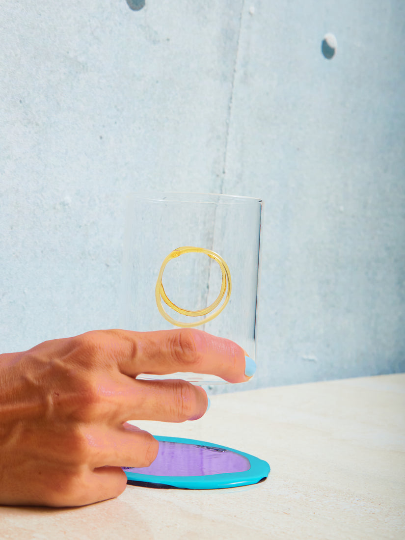A hand holds a clear glass with yellow flame worked circle on opposite sides.