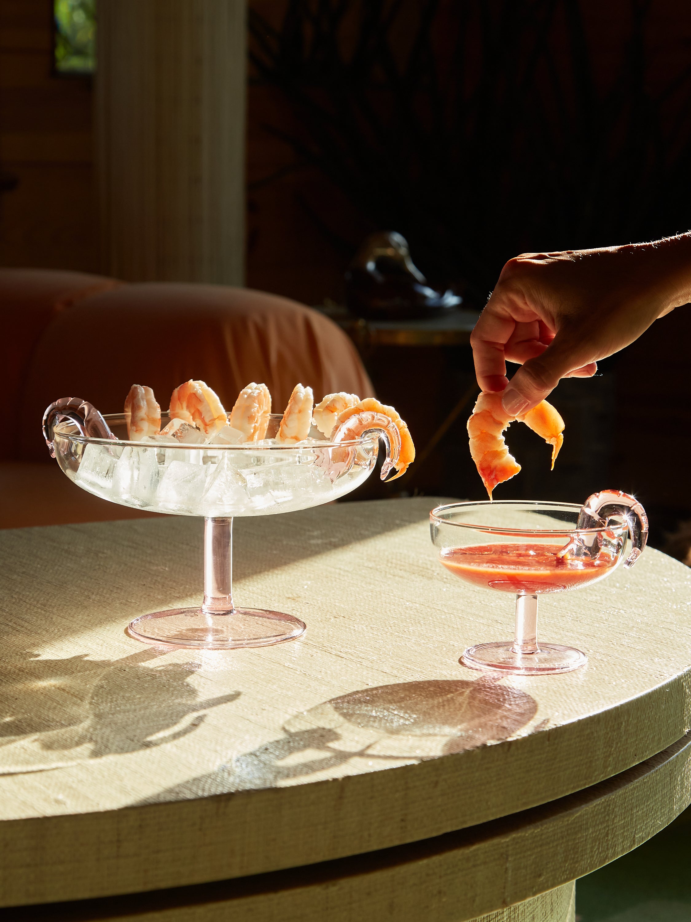 A hand dunking a shrimp into a coupe of cocktail sauce next to a platter full of ice with shrimp alongs its rim.