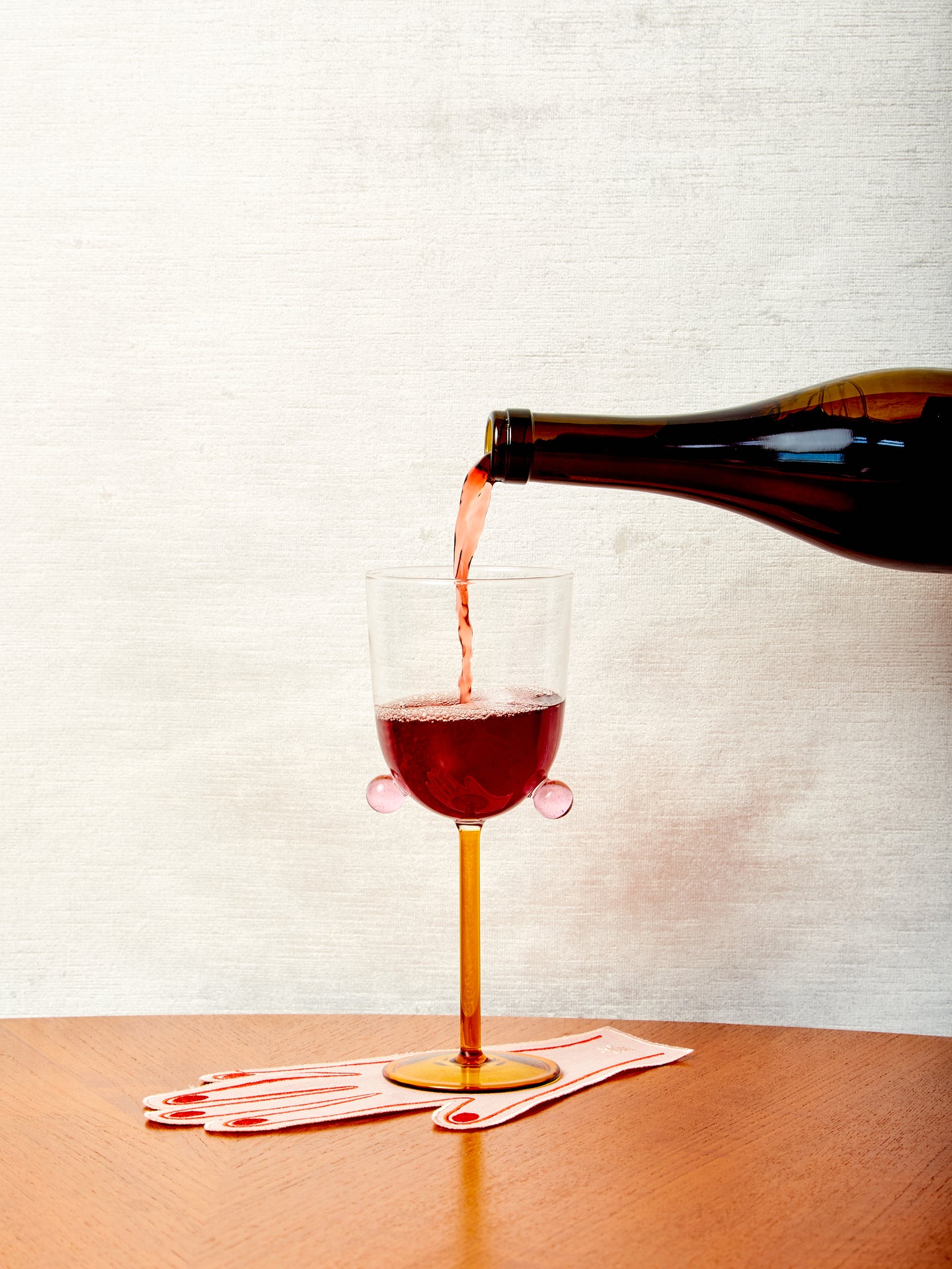 A bottle of red wine pouring into a honey and pink wine glass atop a hand cocktail napkin.