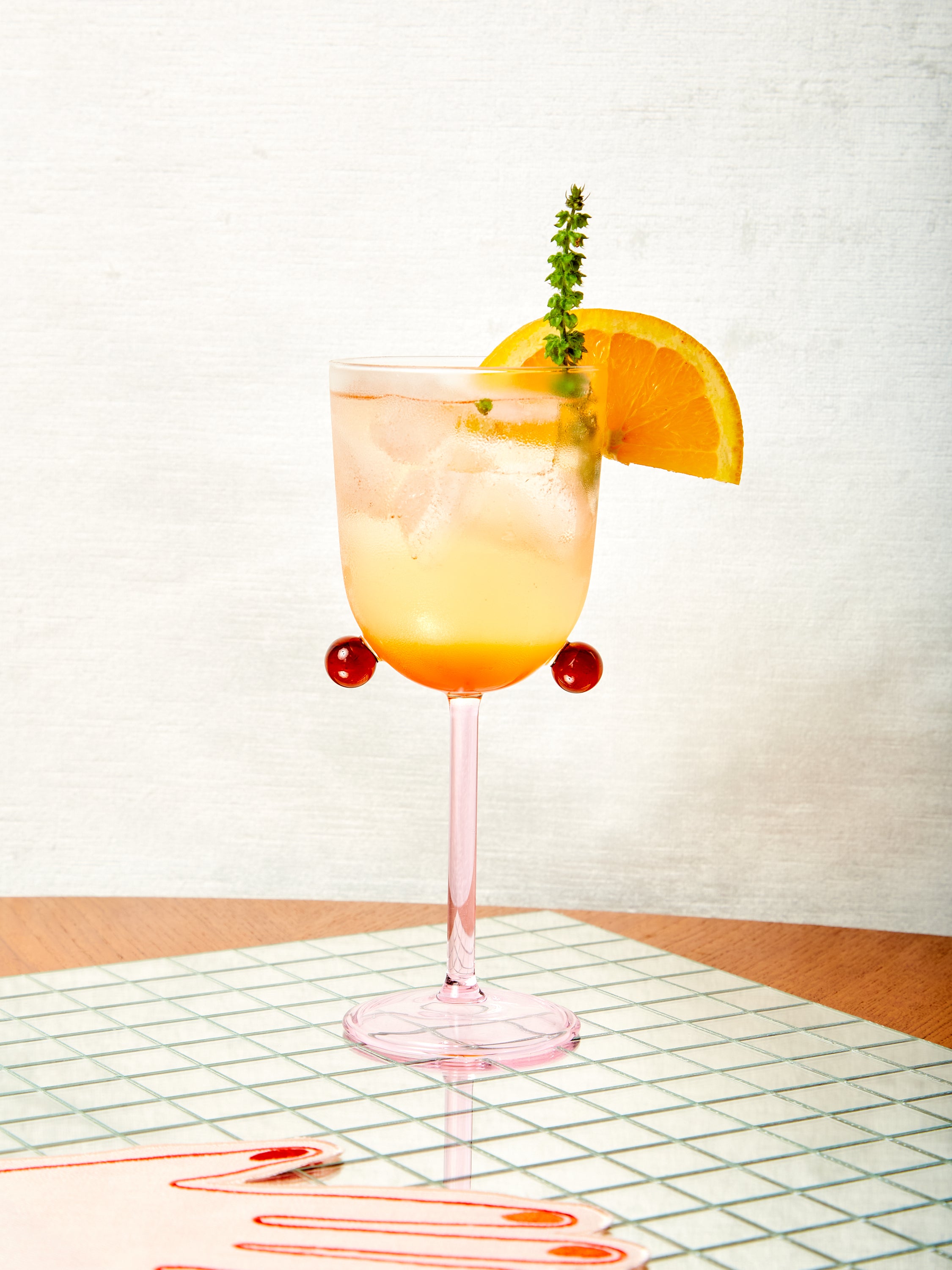 A pink and amber Pompom glass with light amber cocktail, orange wedge, and herb.