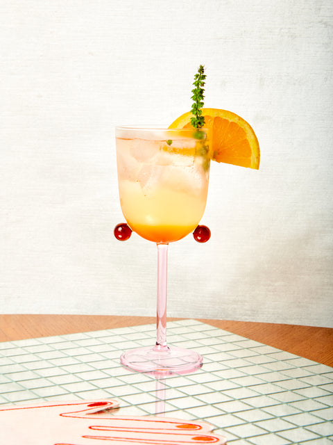 A pink and amber Pompom glass with light amber cocktail, orange wedge, and herb.