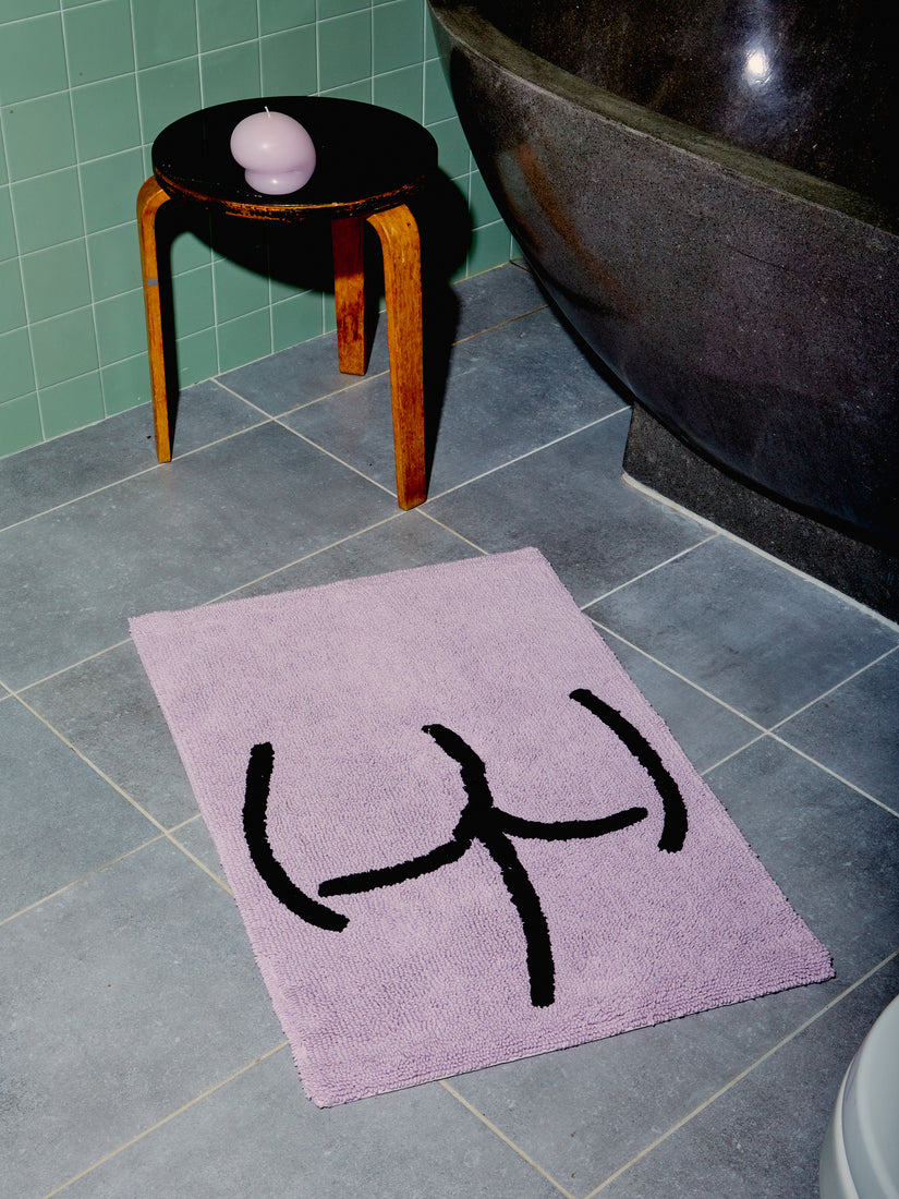 Tushy Bath Mat in Lavender styled in a sage green tiled bathroom with black wooden stool, candle, and stone tub.
