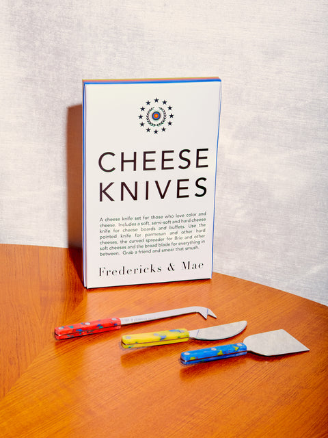 Cheese Knives in three different shapes with red, yellow, and blue handles by Fredericks and Mae. The Cheese Knives sit atop a round wooden table just in front of their packaging.