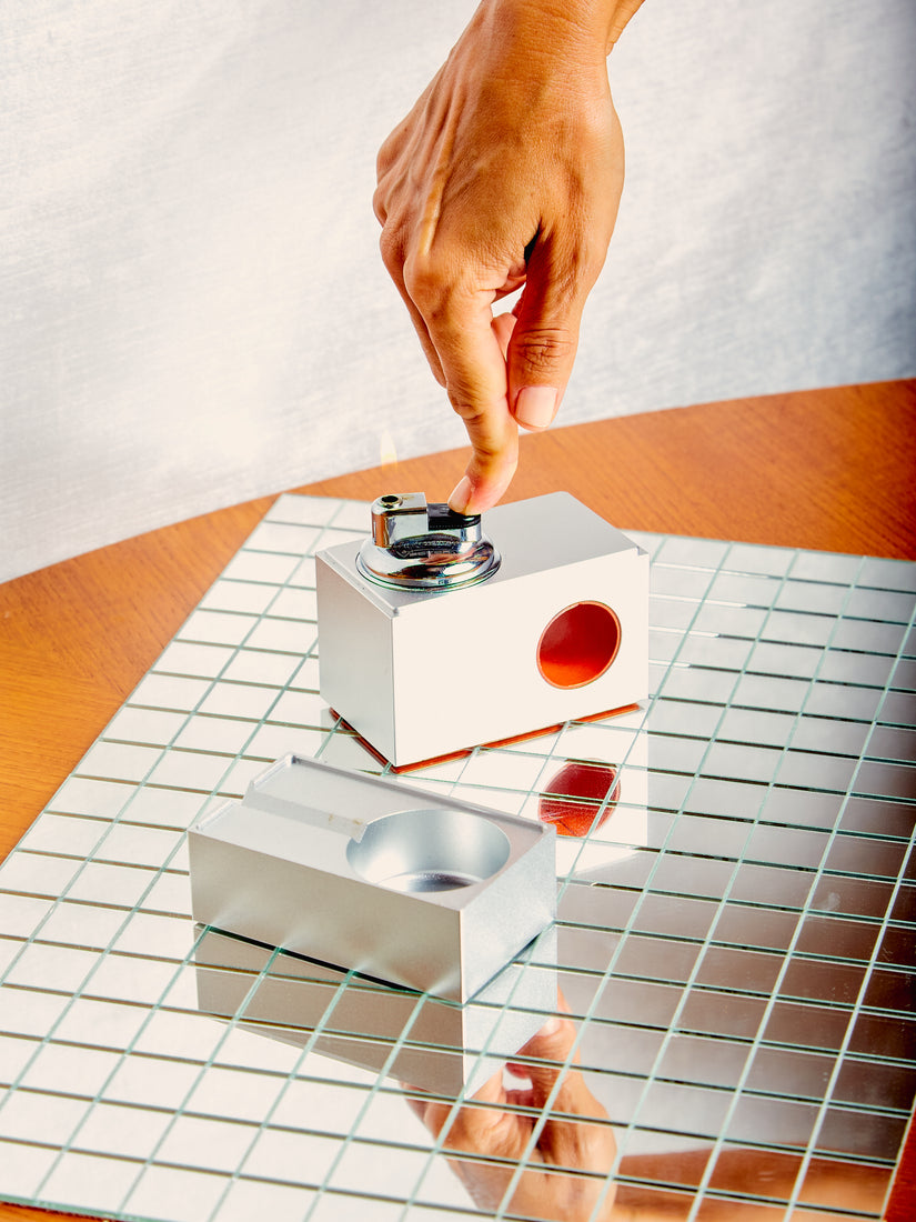 A finger pushes down on the lighter of a Block Table Lighter by Houseplant. The lid/ashtray is featured next to the lighter.