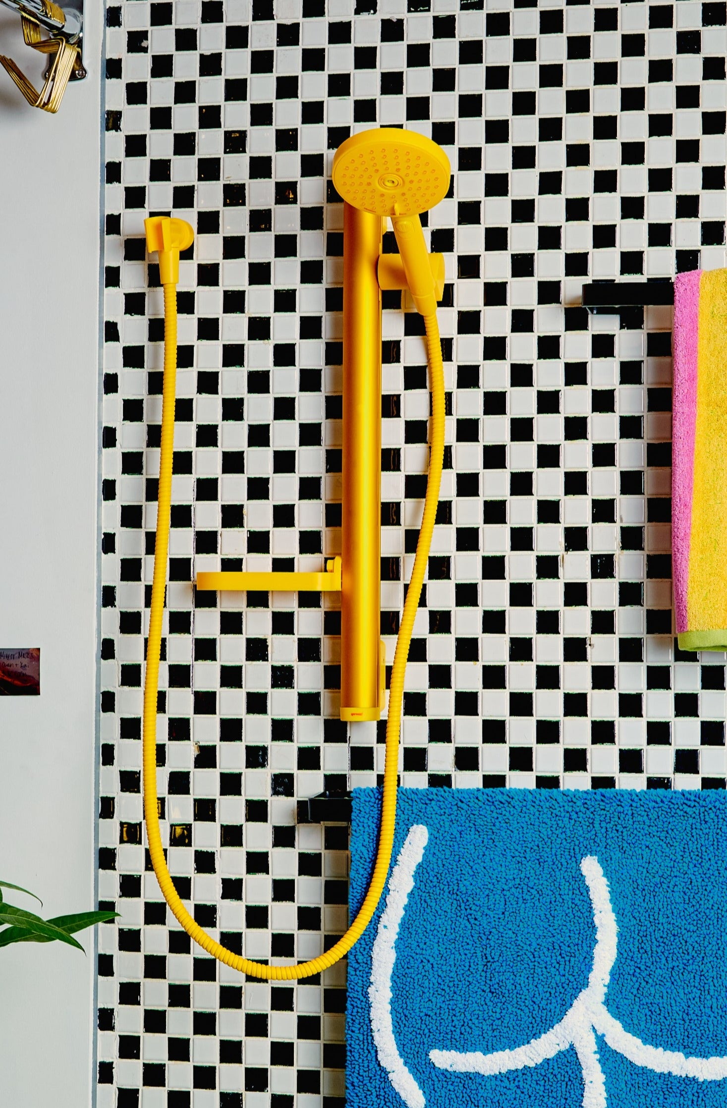 A yellow Minimalist Shower Head installed on a black and white checkered tiled wall besides towel racks.