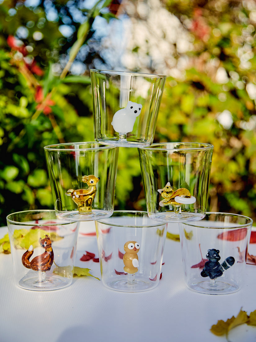 A pyramid of six different Animal Tumblers by Ichendorf Milano sit amongst a loose scattering of leaves on a white table top. In the background, out of focus, is a garden of foliage.