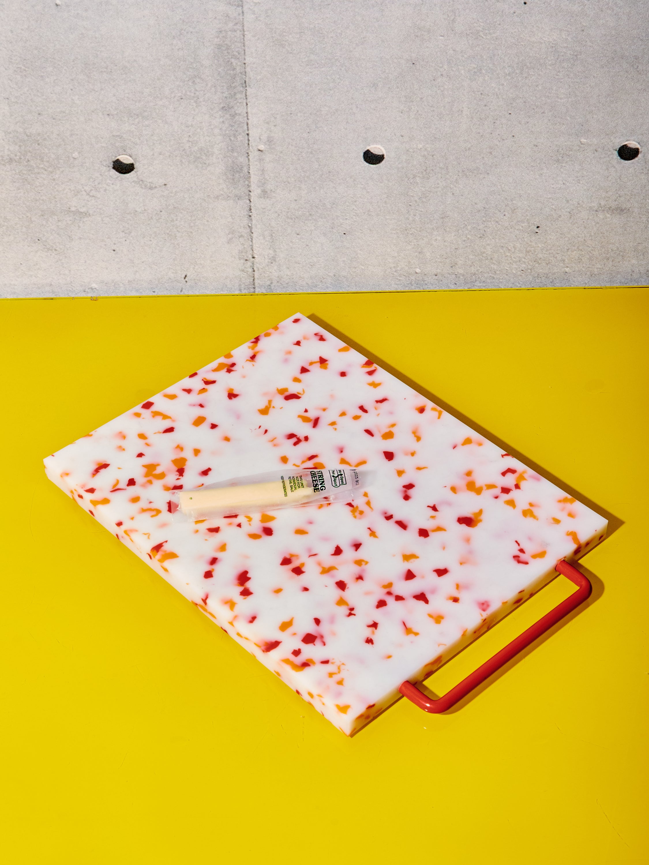 A white chopping block with red and orange speckles and a red handle sits atop a yellow surface. An individually packaged string cheese sit atop the chopping block.