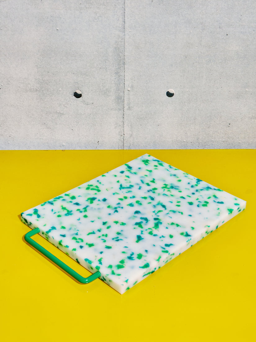 A white chopping block with a green handle, and green speckles of various tones sits atop a yellow surface.