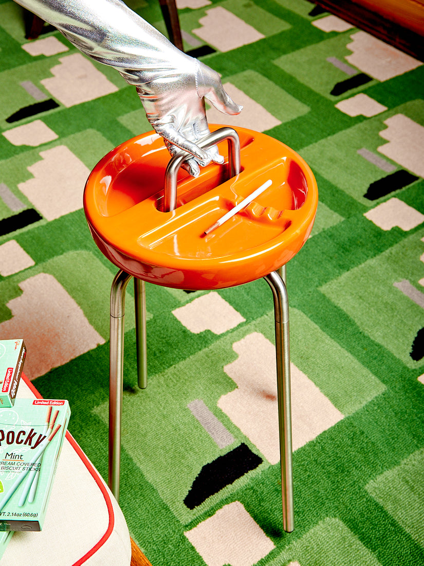 A silver gloved hand holds the metal handle of an orange Side Table Ashtray by Houseplant. The table sits on a green Cold Picnic carpet.