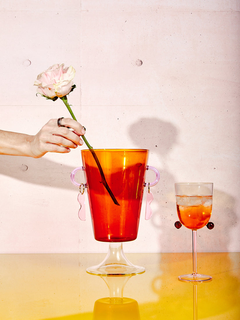 A hand places a single rose into a Happy Vase, just left of a Pompom Wine Glass.