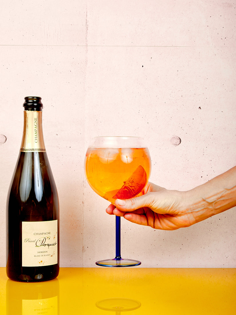 A hand holds up a full Spritz Glass to the right of a bottle of champagne.