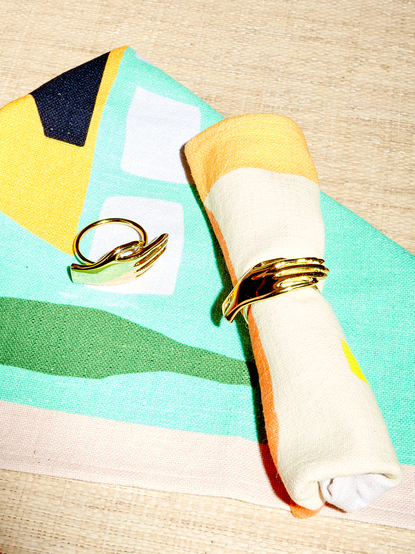 A pair of Dada napkin rings are displayed with abstract dinner napkins by Cold Picnic. One napkin rings wraps around a rolled napkin while the other sits atop a folded one.
