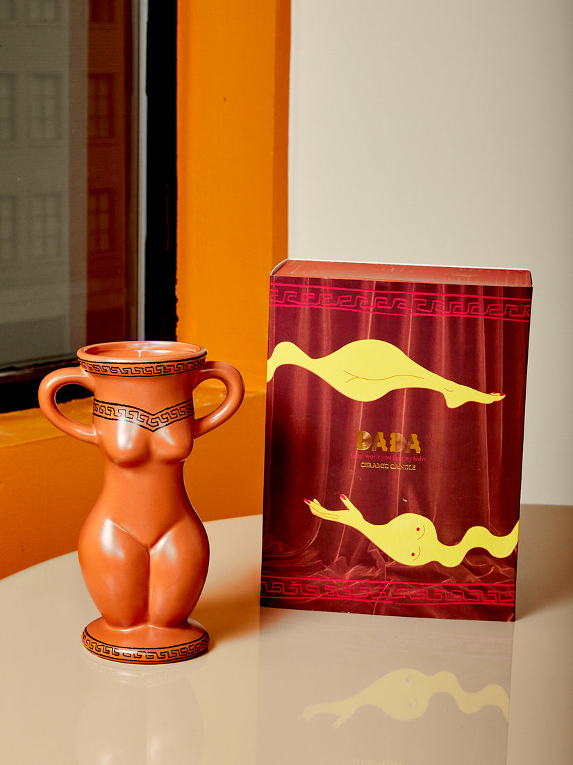 A ceramic vessel in the form of a feminine body that is also a candle. The candle sits next to the box it comes packaged in.