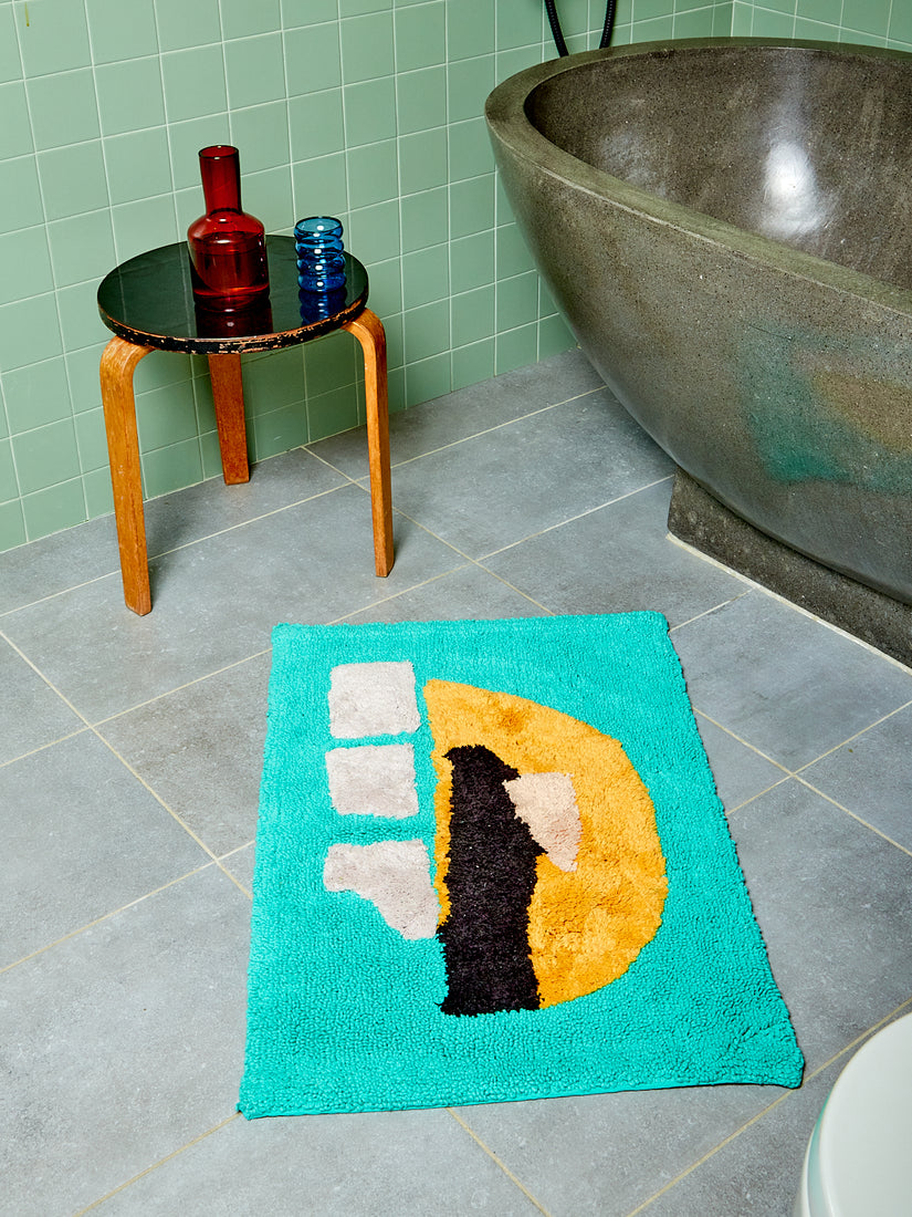 Bright teal bathmat with tangerine, cream, and black abstract accents by Cold Picnic in a sage tiled bathroom with wooden side stool, stone tub, amber carafe, and blue glass.