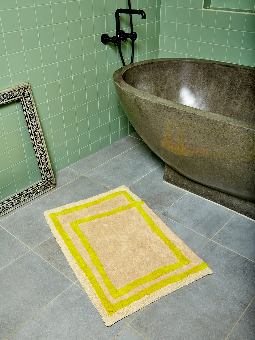 The Frames Bath Mat by Cold Picnic sits in a sage green tiled bathroom in front of a rounded stone bathtub.