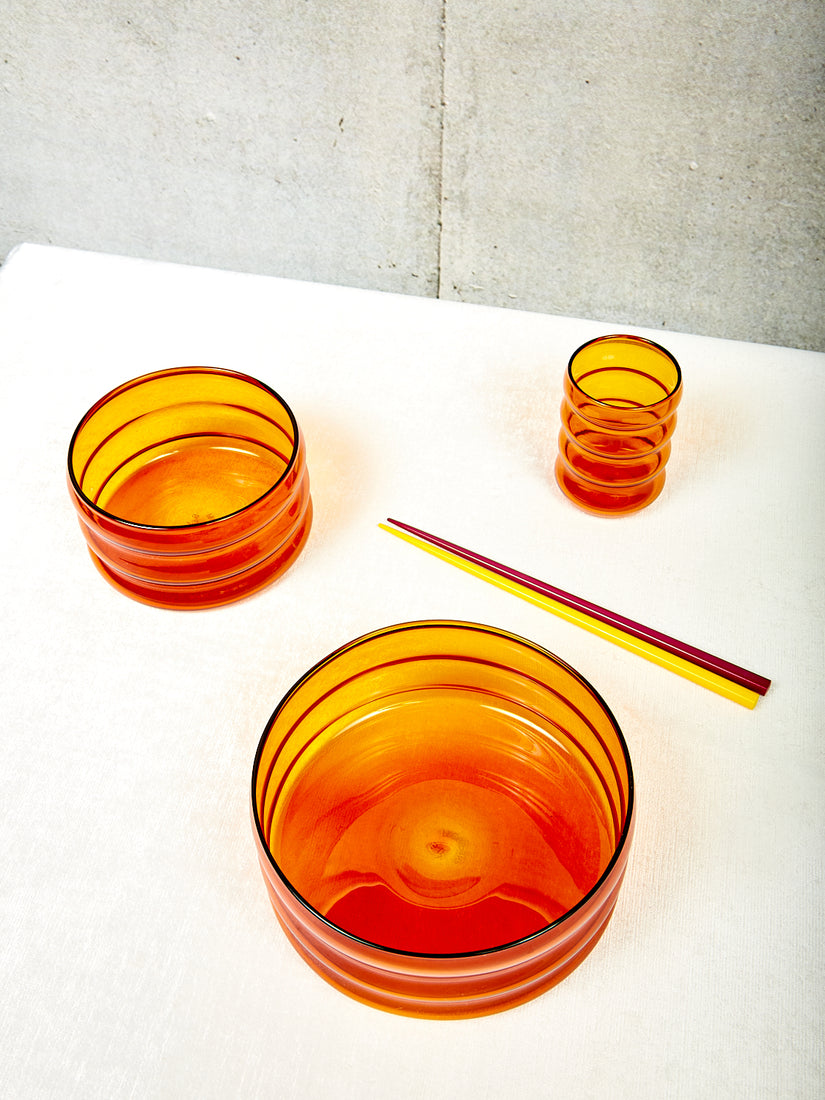 A pair of yellow and burgundy chopsticks sits amongst an amber ripple cup, and small and medium amber ripple bowls.