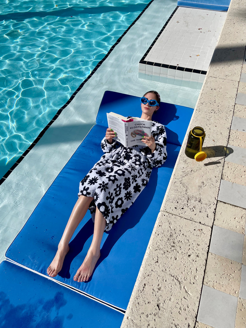 Madde lounges on a pool float, reading a book, wearing a black and white robe.