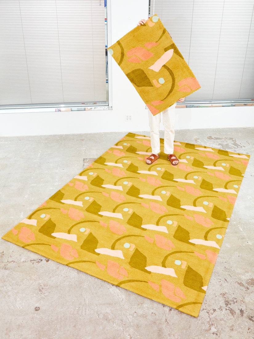 Someone holds a 2x3 The Aviary, Late Summer rug in front of their body while standing on a 4x6 version of the rug.